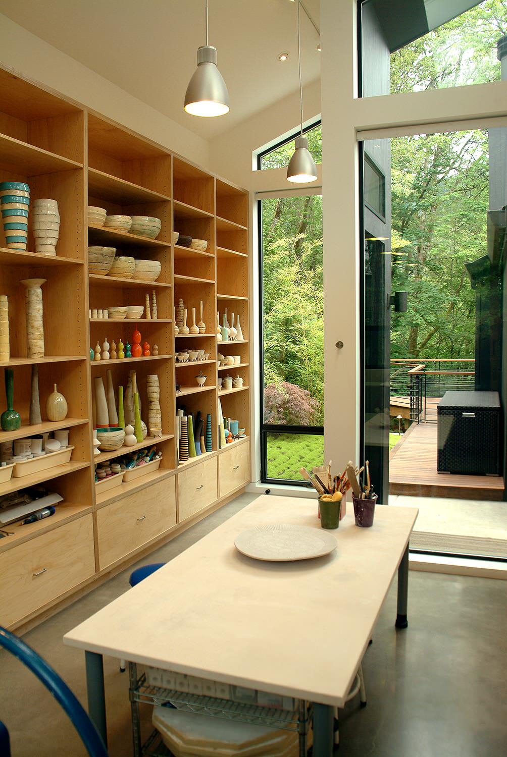 Marika VanAdelsberg's ceramic studio with floor-to-ceiling wooden shelving displaying pottery and ceramics, large windows overlooking lush greenery