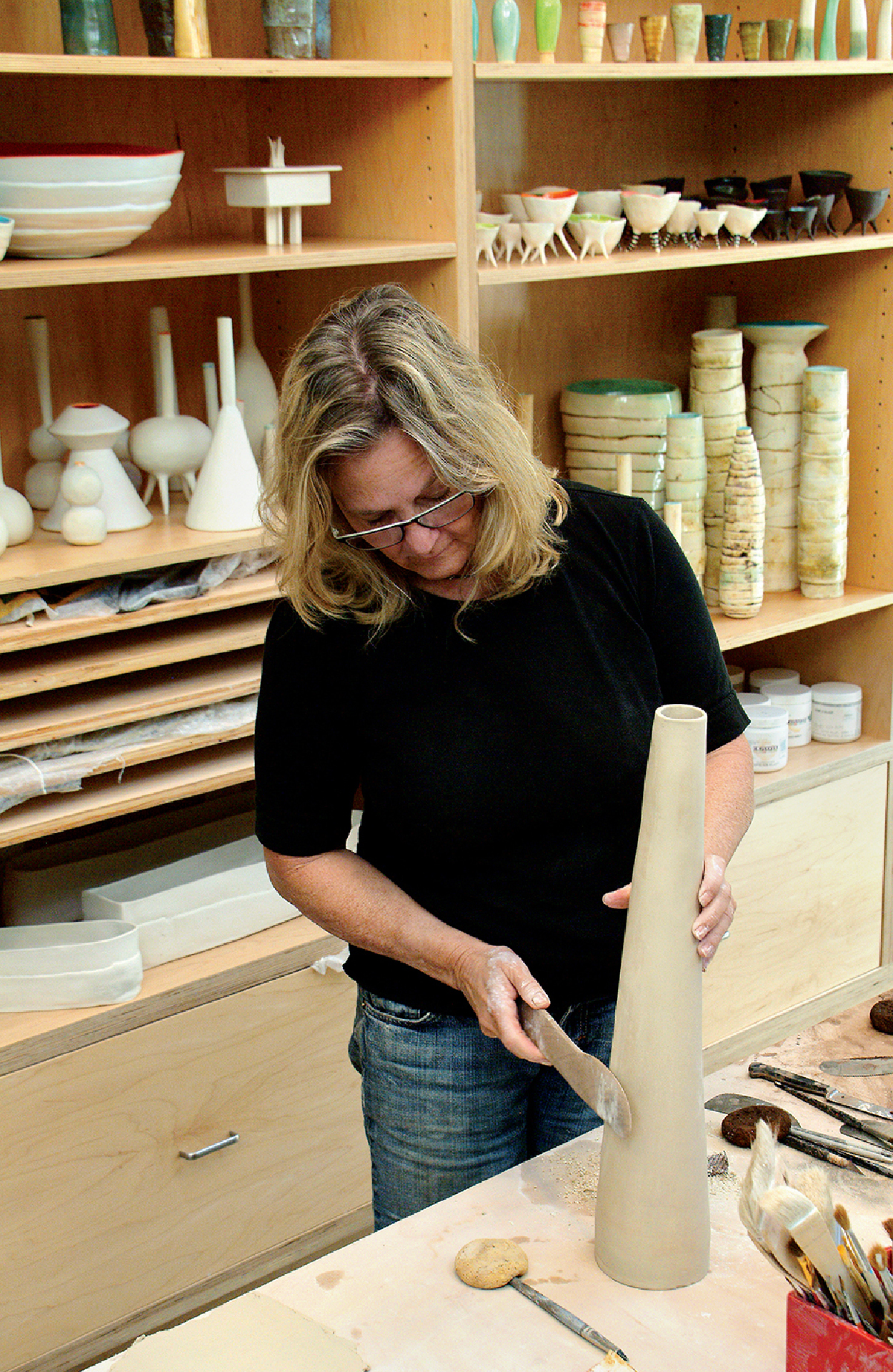 Marika VanAdelsberg working at her pottery wheel, shaping a tall ceramic vessel in her studio surrounded by shelves of finished pottery